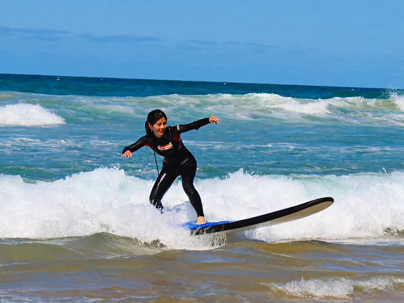 Woman learning to surf, balancing on a surfboard while riding a small wave near the shore.