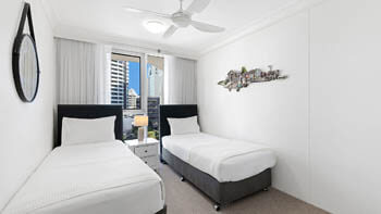 Second bedroom at ULTIQA Beach Haven on Broadbeach with two single beds, bedside table and lamp, and a window overlooking nearby high-rises.