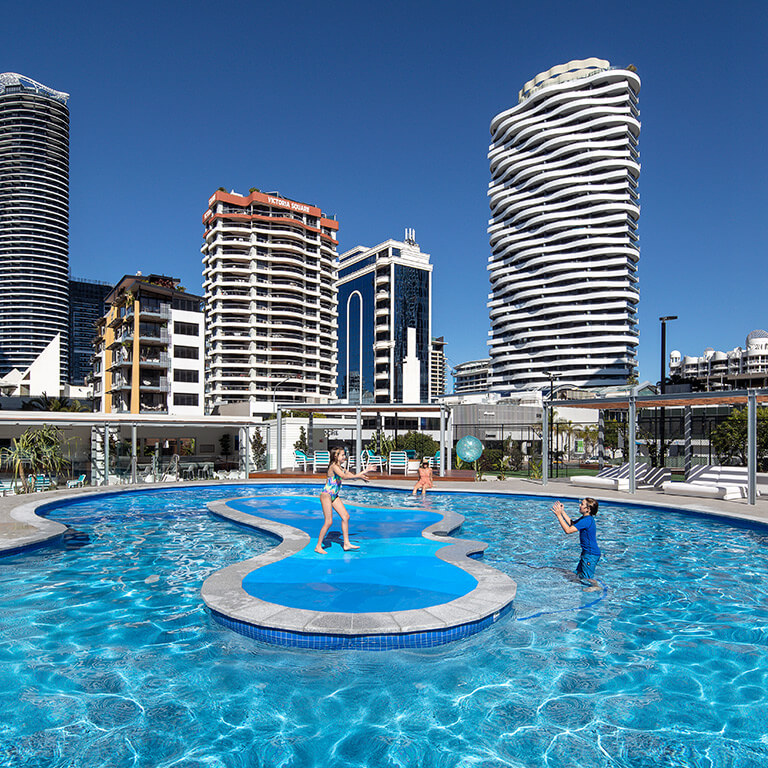 Couple relaxing on poolside loungers, with a woman smiling while reading a book and a man sitting nearby.