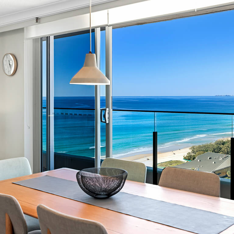 Bedroom with large windows at ULTIQA Points North Coolangatta offering panoramic ocean and coastline views, featuring a neatly made bed with soft pink cushions.